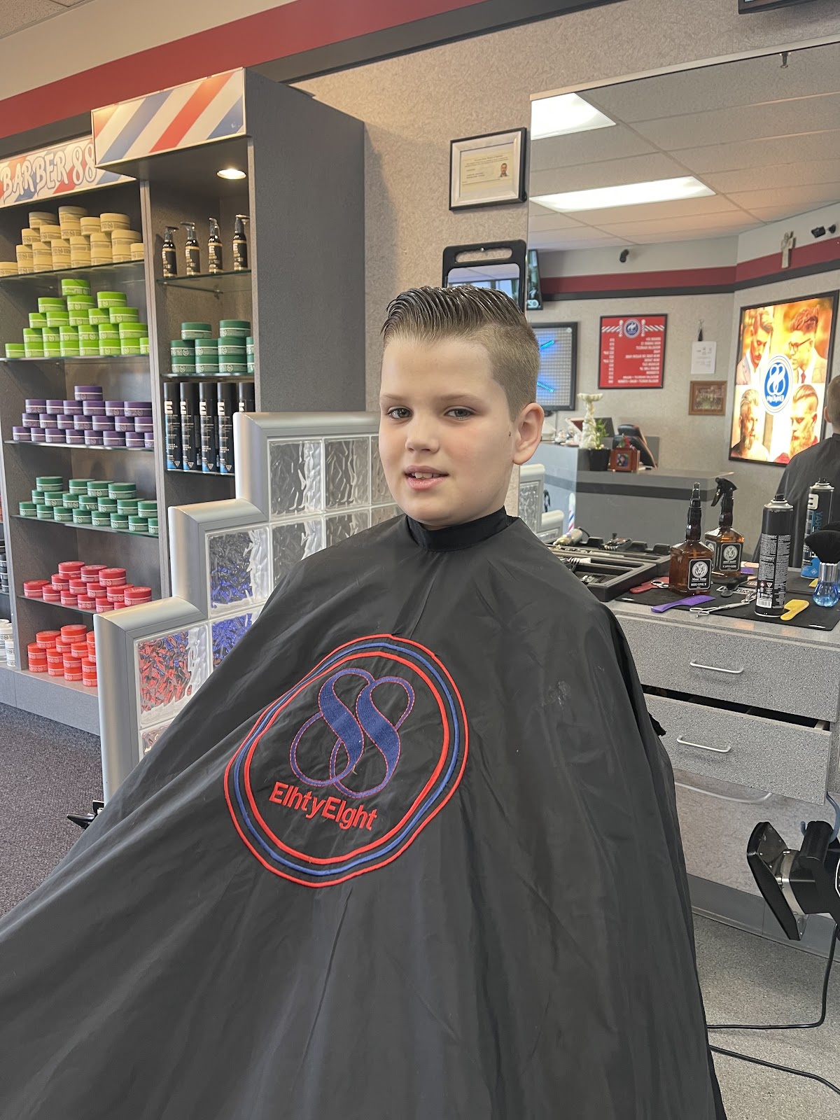A boy with a fresh side-part fade sits in our black 88 cape in front of the pomade shelf