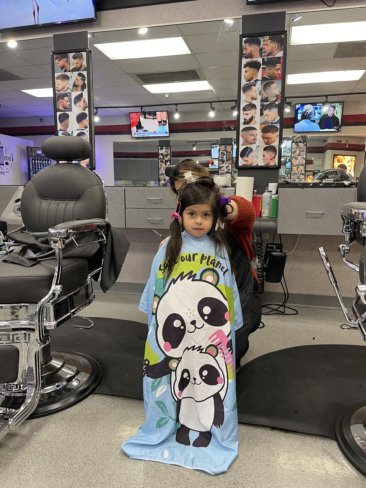 A little girl with clips in her pigtails wears a blue panda cape while getting a trim at Barber 88