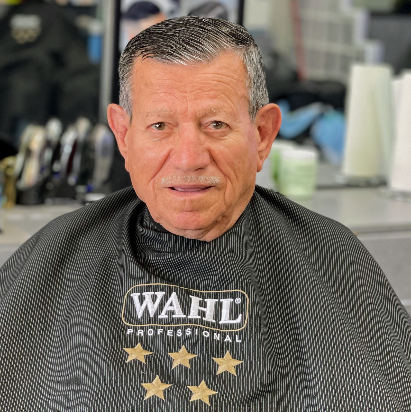 A silver-haired gentleman with a fresh cut and neat mustache smiles softly in a pinstripe cape