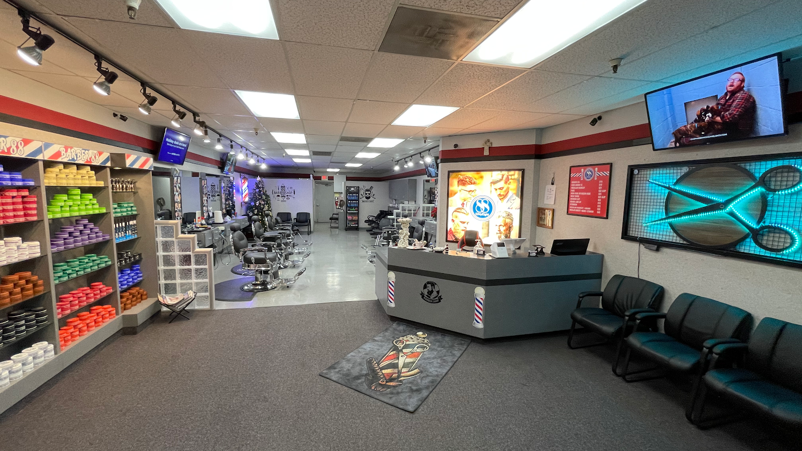 Inside Barber 88: a shelf wall stacked with colorful pomade tubs, a row of barber chairs down the gray floor, and the lit 88 portrait sign over the front desk